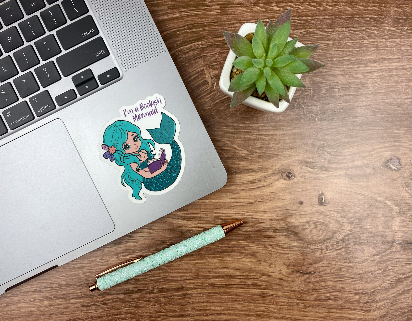 Reading mermaid sticker applied to the corner of a silver laptop trackpad, next to a succulent and a glitter pen on a wood desk