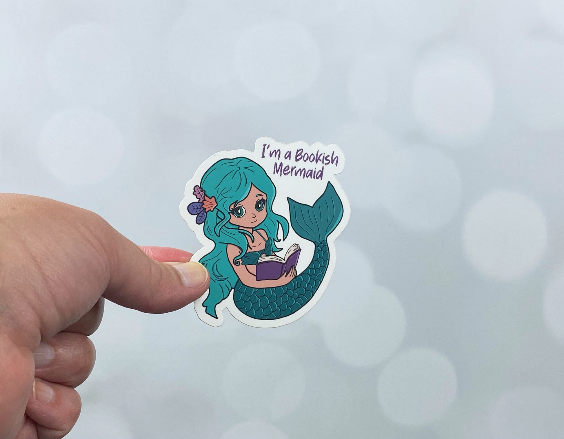 Die-cut vinyl sticker held between fingers, showing a teal-haired mermaid reading a book, with the text "I'm a bookish mermaid," on a light bokeh background