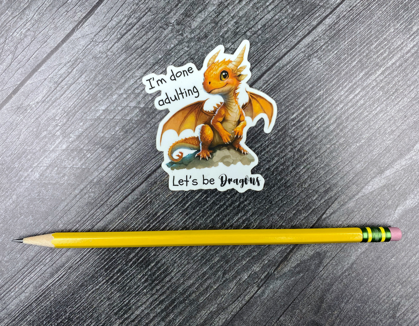 Die-cut vinyl sticker showing an orange baby dragon perched on a rock, with the text "I'm done adulting, let's be dragons," on a gray wood background next to a yellow pencil