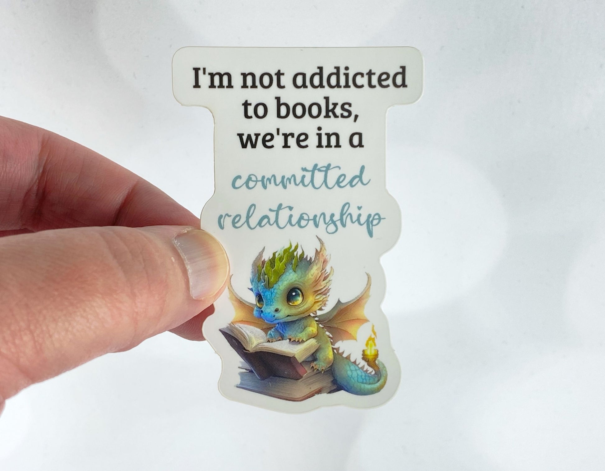 A hand holds a sticker illustration of a baby dragon reading an open book with the phrase "I'm not addicted to books, we're in a committed relationship," against a white background.