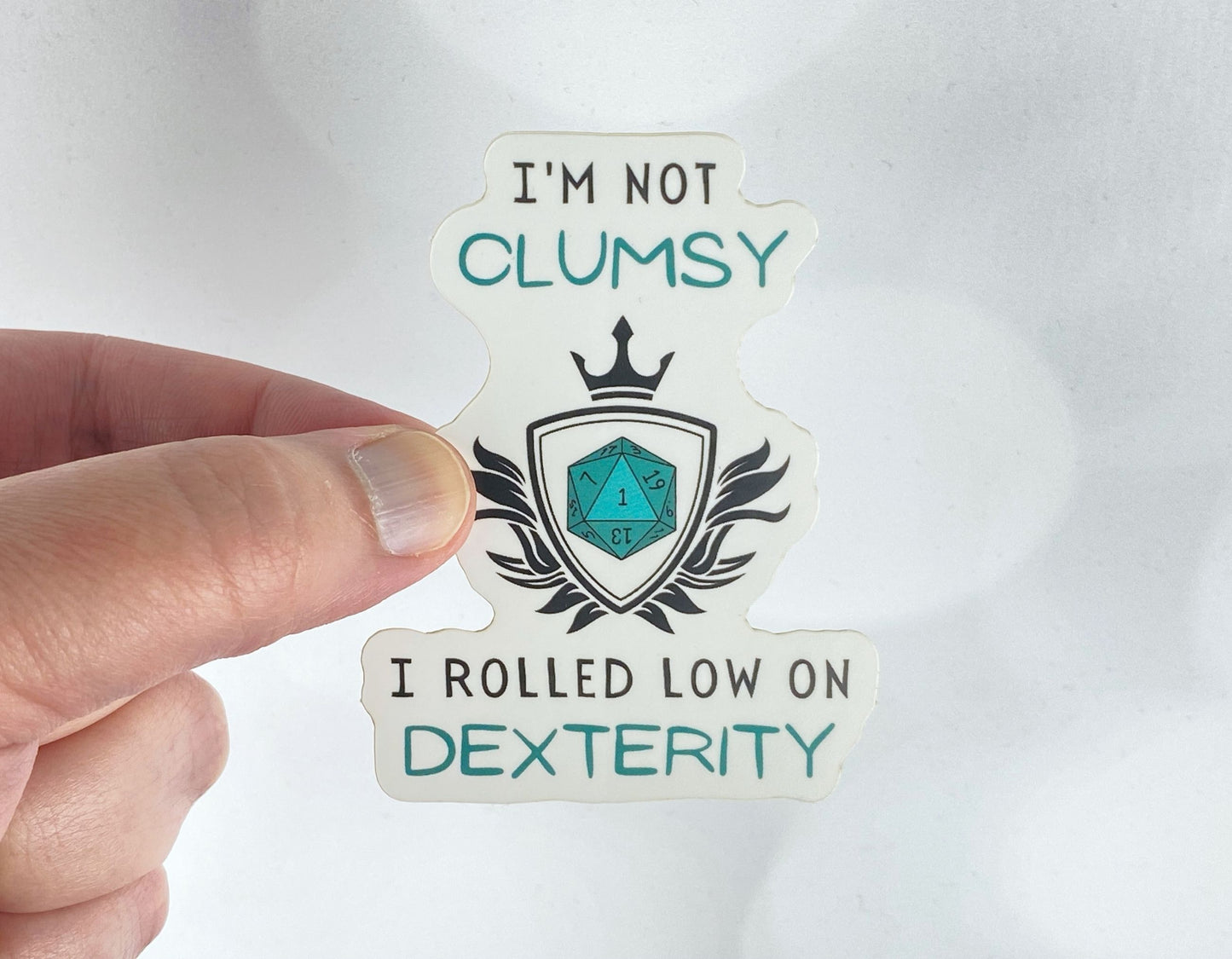 A hand holds a sticker reading "I'm not clumsy, I rolled low on Dexterity" with a crowned shield and a teal twenty-sided die showing the number 1, against a white background.