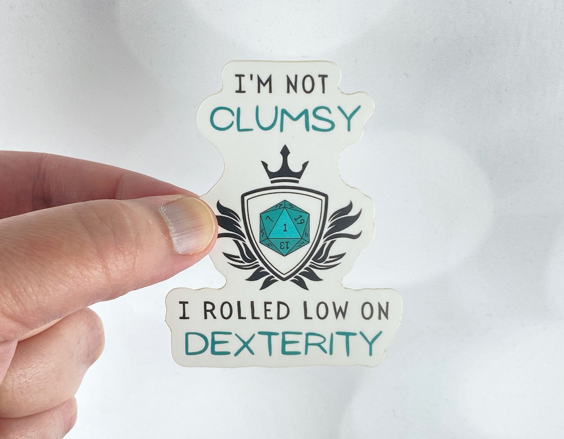 A hand holds a sticker reading "I'm not clumsy, I rolled low on Dexterity" with a crowned shield and a teal twenty-sided die showing the number 1, against a white background.