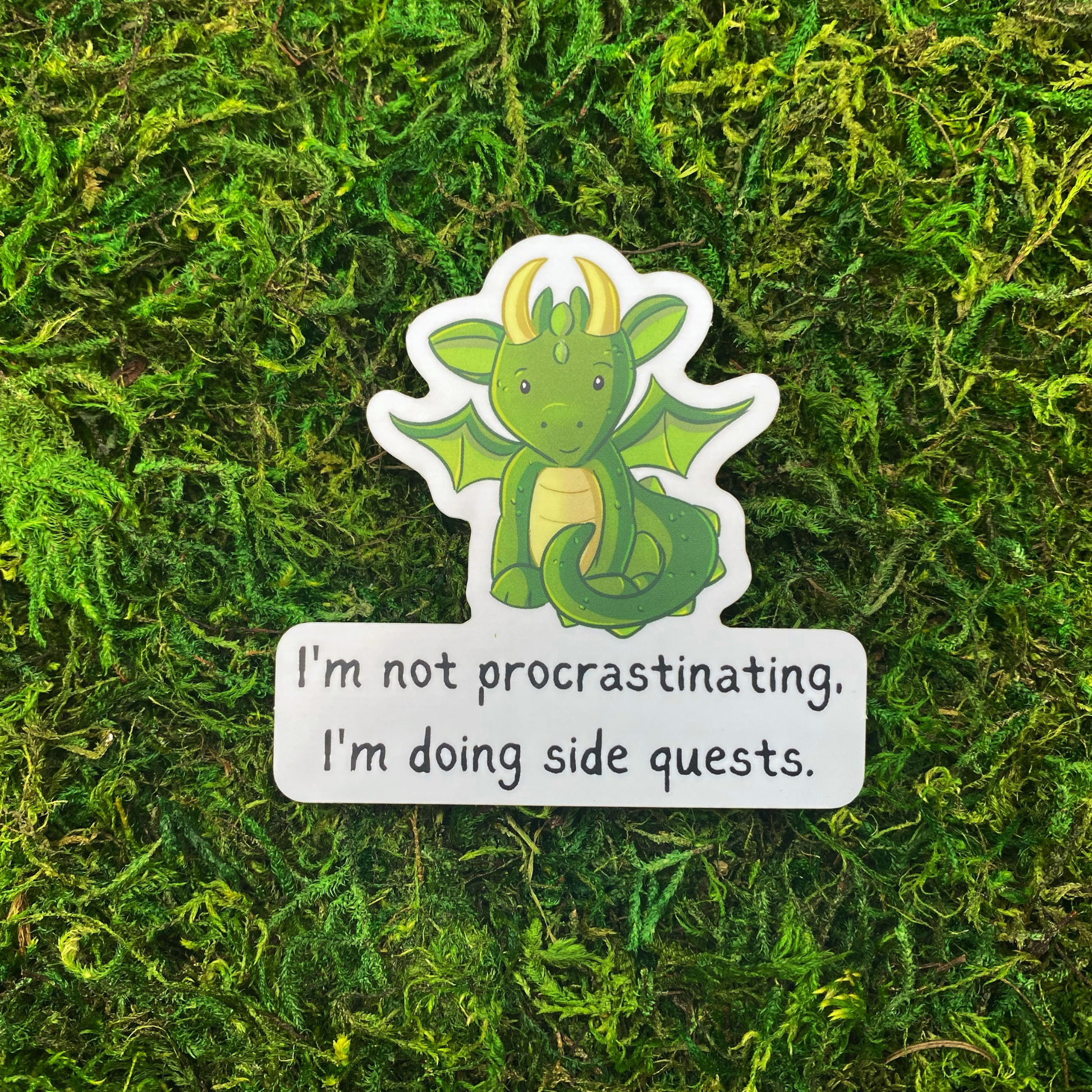 A sticker with an illustration of a green baby dragon above the text "I'm not procrastinating, I'm doing side quests," resting on artificial moss.