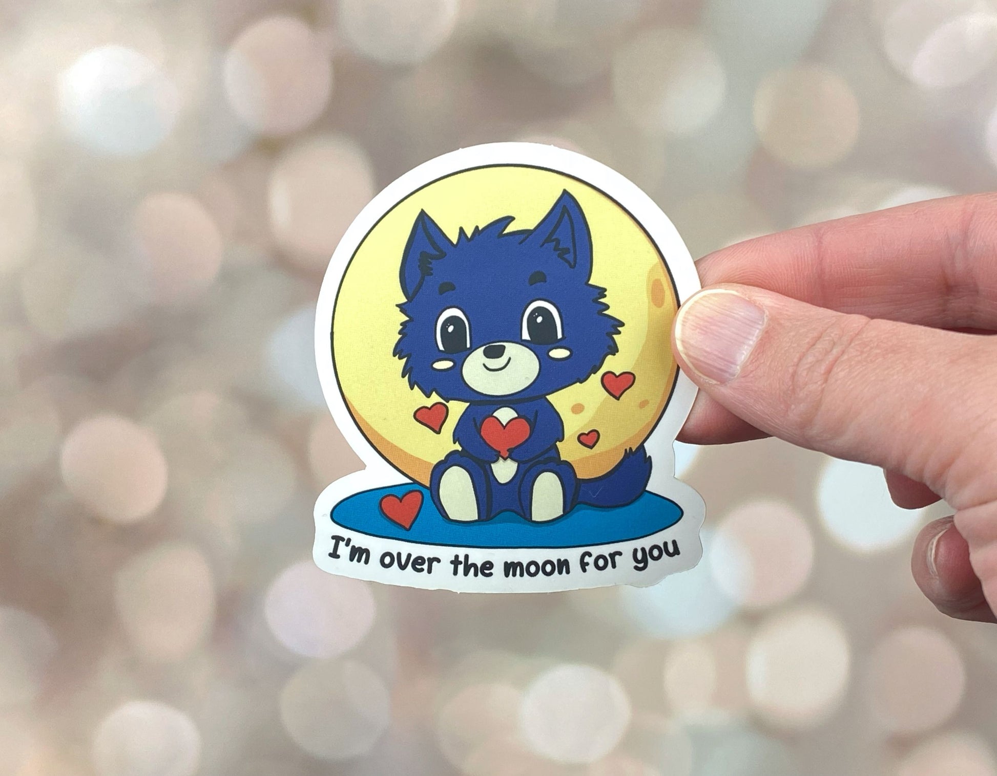 Die-cut vinyl sticker showing a grey werewolf pup holding a heart in front of a full moon, with the text "I'm over the moon for you," on a golden bokeh background