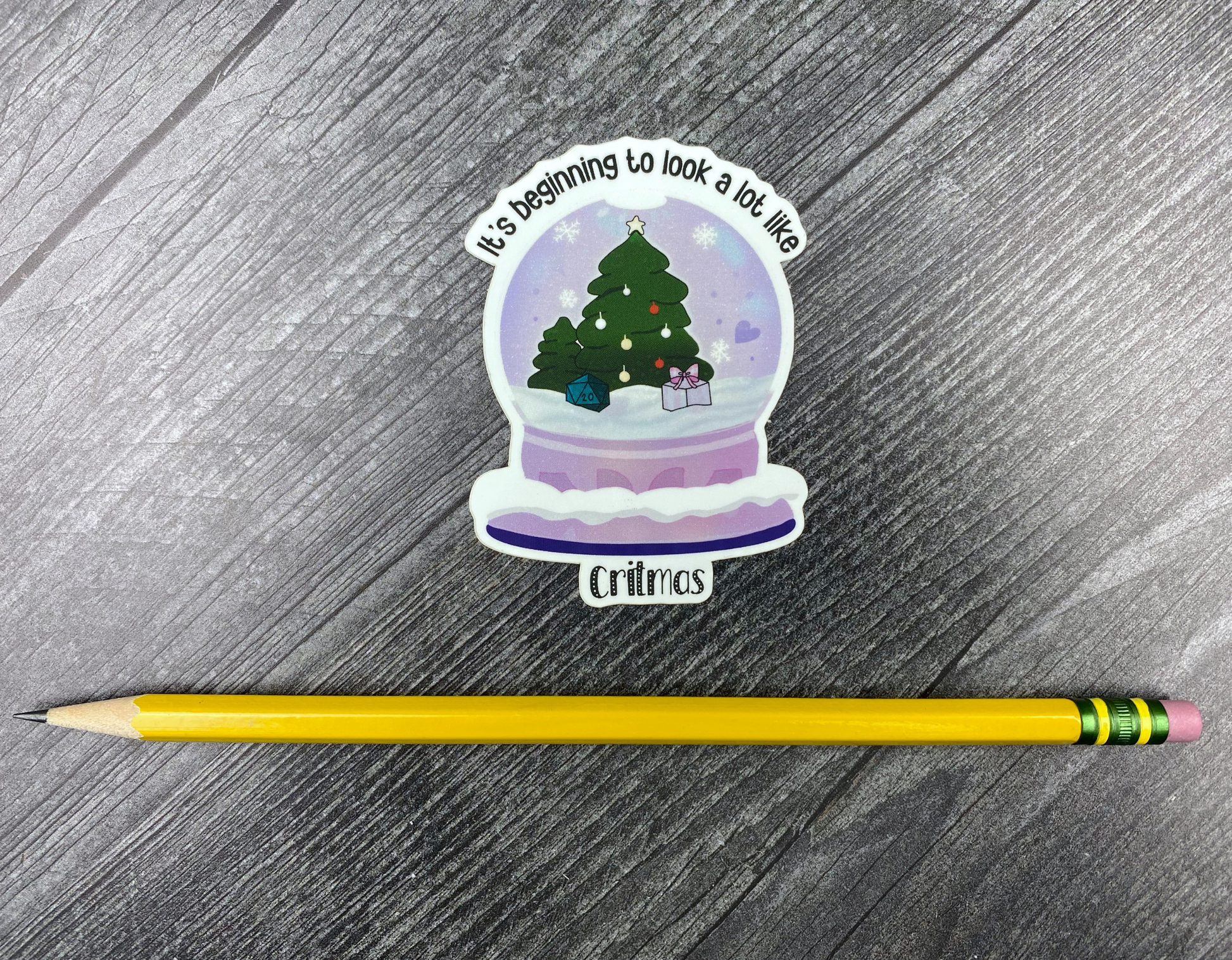 Die-cut vinyl sticker showing a snow globe with a Christmas tree, a d20, and a wrapped gift, with the text "It's beginning to look a lot like Critmas," on a gray wood background next to a yellow pencil