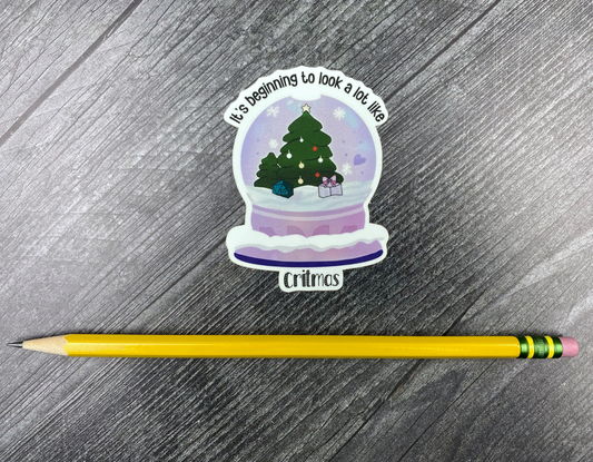 Die-cut vinyl sticker showing a snow globe with a Christmas tree, a d20, and a wrapped gift, with the text "It's beginning to look a lot like Critmas," on a gray wood background next to a yellow pencil