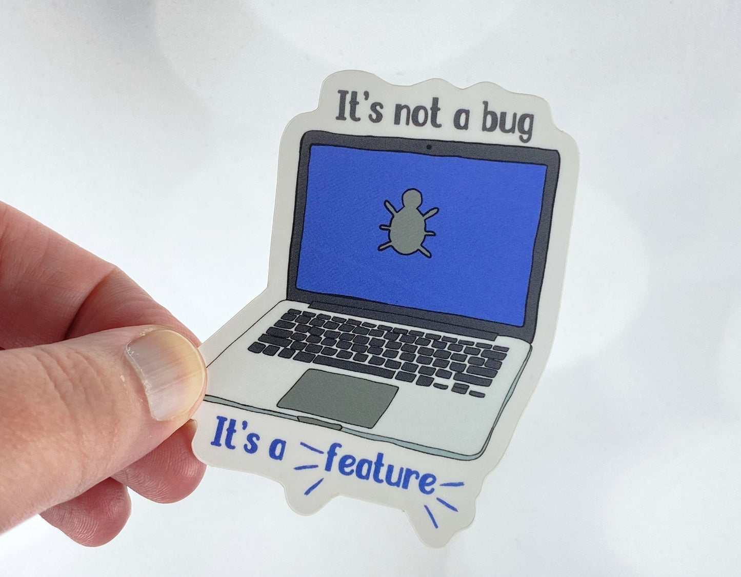 Die-cut vinyl sticker held between fingers, showing a laptop with a bug icon on the screen, with the text "It's not a bug, it's a feature," on a white background