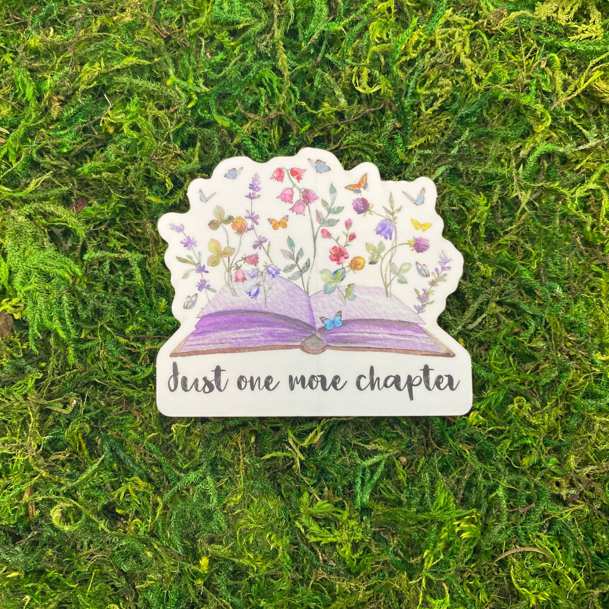 A sticker illustration of an open book with wildflowers and butterflies blooming from its pages, reading "Just one more chapter," resting on artificial moss.