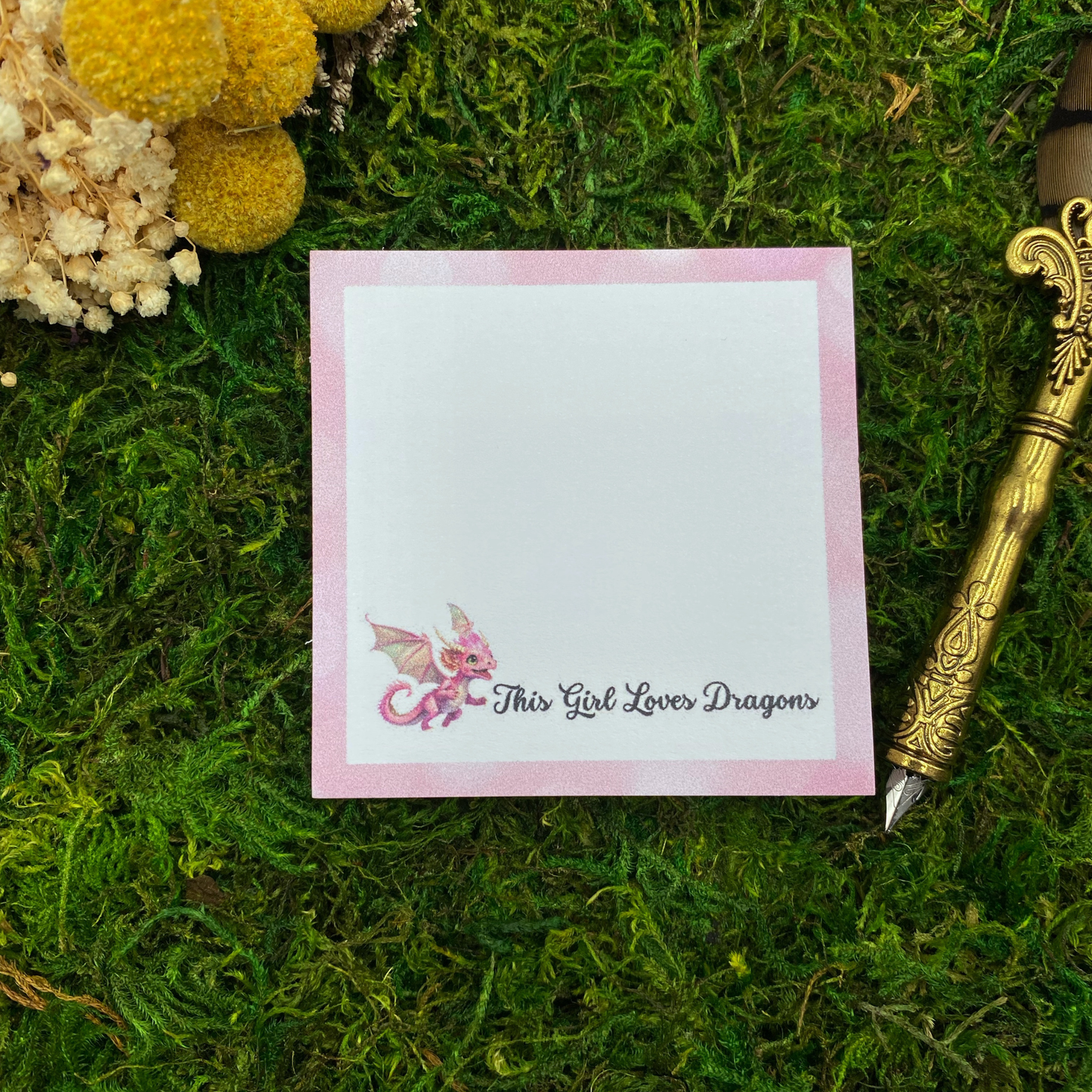 Square sticky note pad with a pink border, a small kawaii pink dragon illustration, and the words "This Girl Loves Dragons," resting on moss next to dried yellow and white flowers and a gold quill pen.