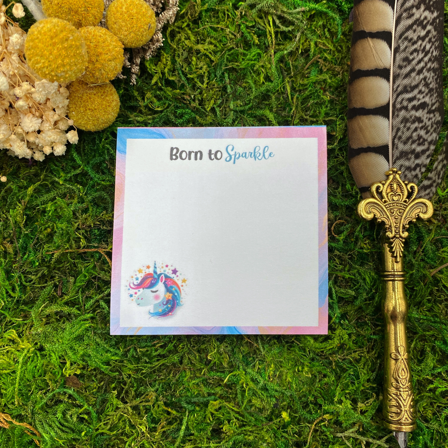 Square sticky note pad with a pastel marbled border, a rainbow-maned unicorn illustration surrounded by stars, and the words "Born to Sparkle," resting on moss next to dried yellow and white flowers and a gold quill pen.