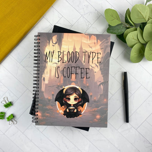 Spiral-bound notebook with kawaii vampire girl illustration and 'My Blood Type Is Coffee' text, styled with a yellow cloth and greenery
