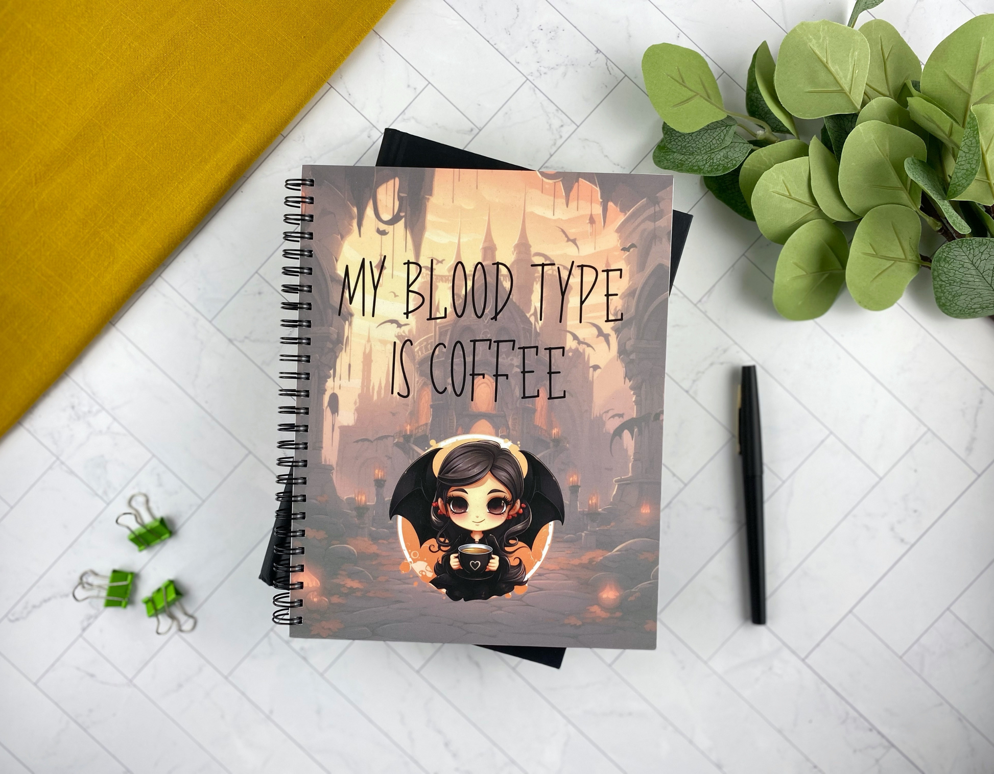 Notebook cover illustrated with a chibi vampire girl with bat wings holding a coffee cup in a gothic city at dusk with flying bats, reading "My blood type is coffee," resting on a black book with green binder clips and a black pen nearby.