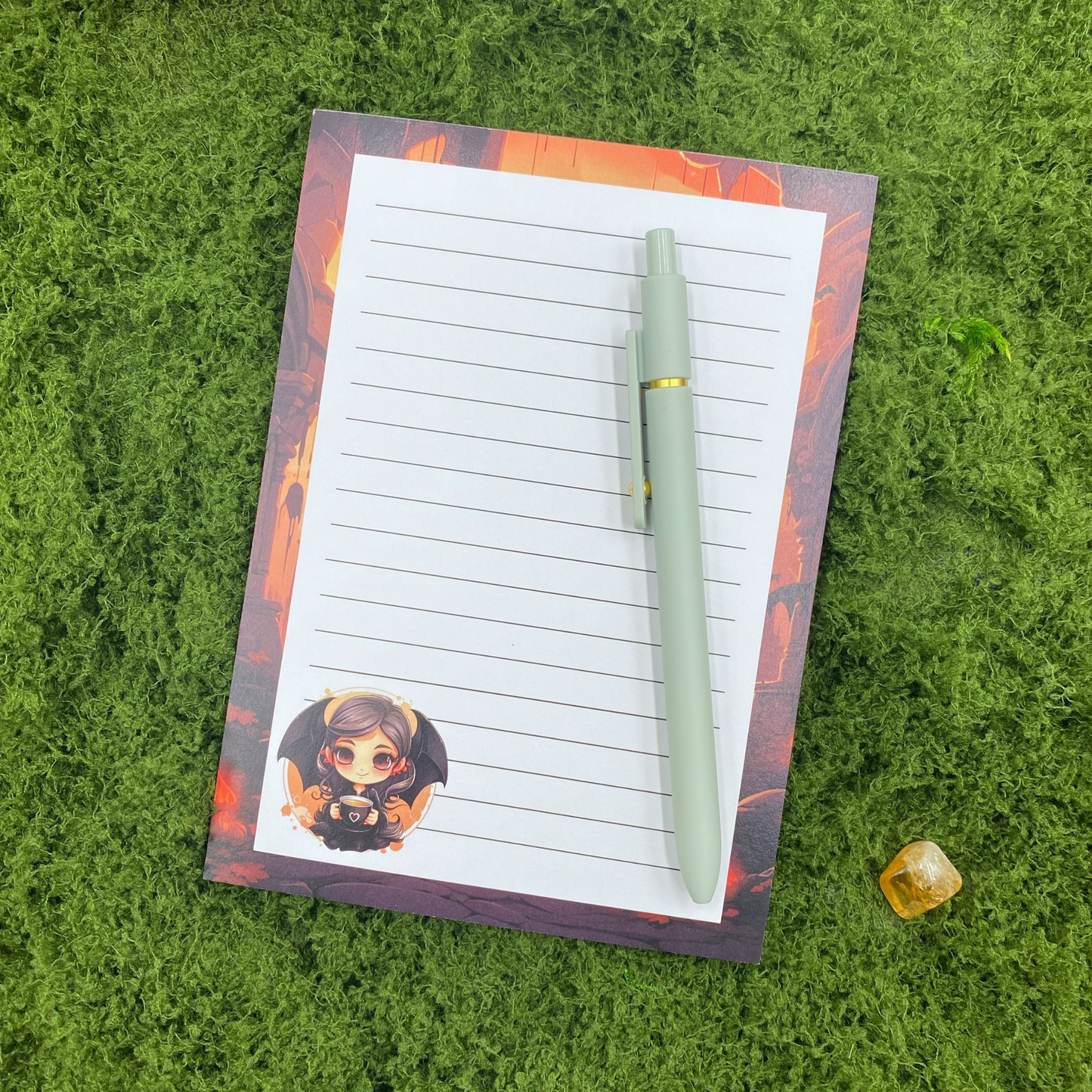 Lined notepad with a gothic castle border in orange and black, and a chibi vampire girl holding a coffee cup in the corner, resting on artificial grass next to a sage green pen and a citrine crystal.