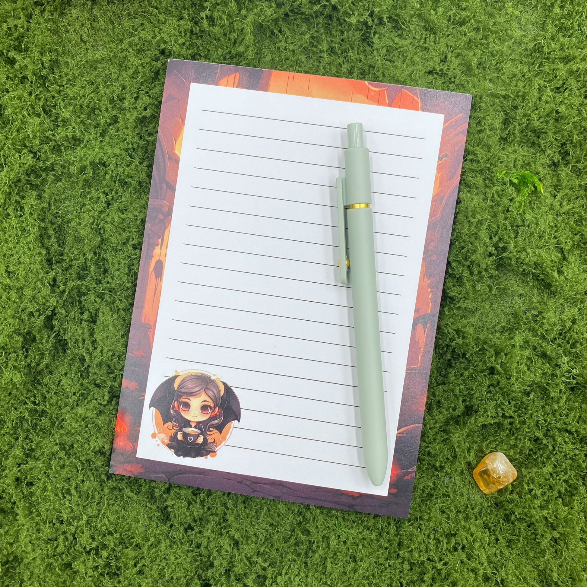 Lined notepad with a gothic castle border in orange and black, and a chibi vampire girl holding a coffee cup in the corner, resting on artificial grass next to a sage green pen and a citrine crystal.