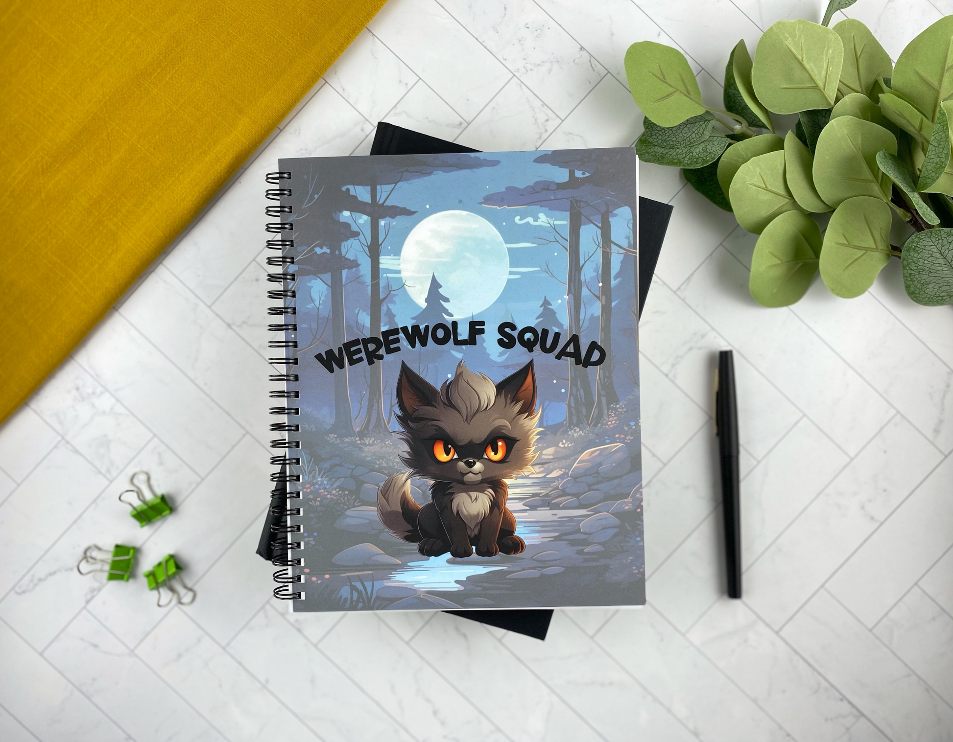 Notebook cover illustrated with a small orange-eyed werewolf cub sitting by a stream in a moonlit forest, with the words "Werewolf Squad," resting on a black book with green binder clips and a black pen nearby.