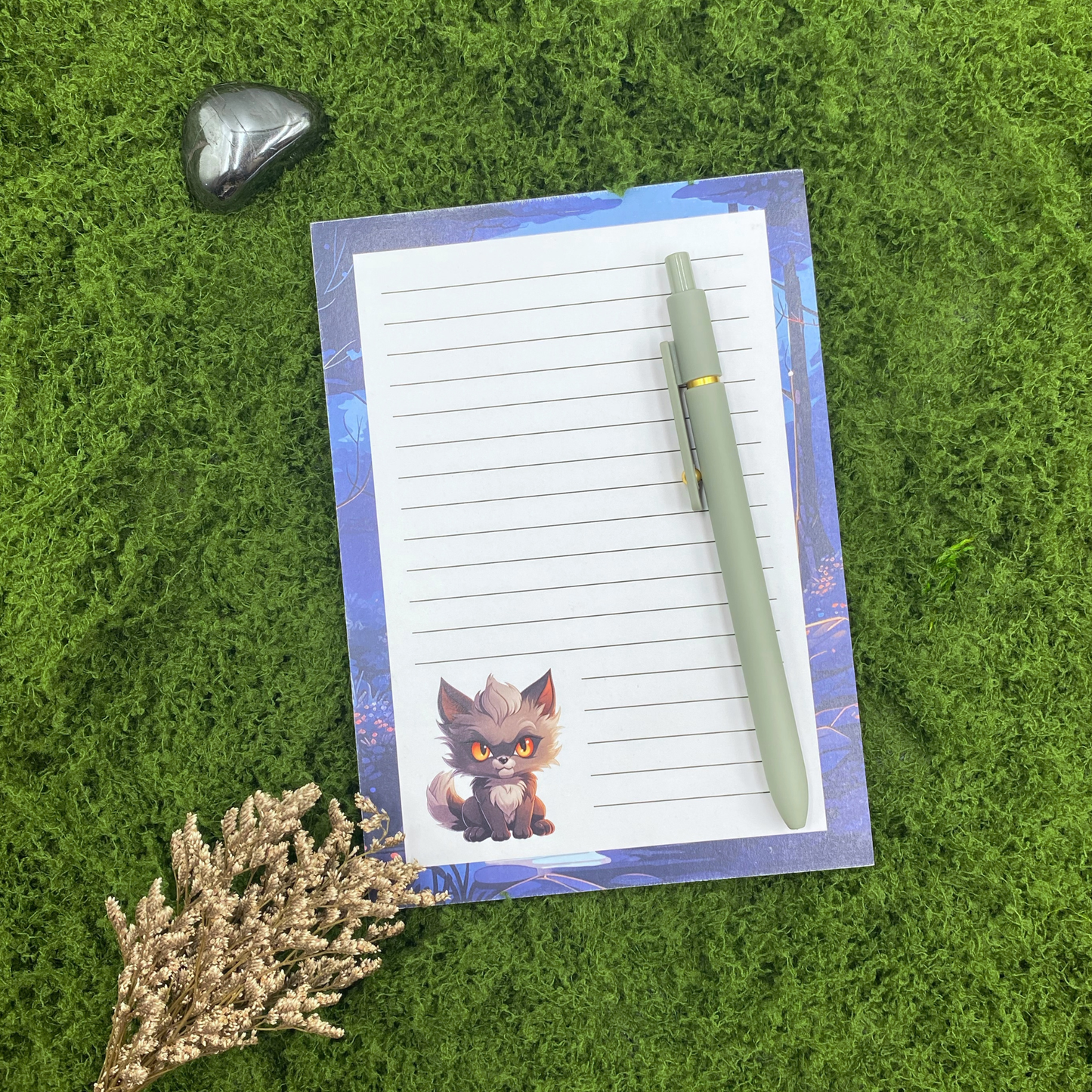 Lined notepad with a moonlit blue forest border and a small orange-eyed werewolf cub illustration in the corner, resting on artificial grass next to a sage green pen, an obsidian stone, and dried flowers.