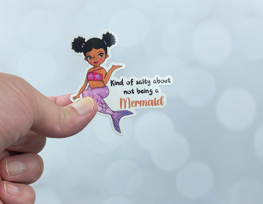 A hand holding a glossy mermaid die-cut sticker featuring a mermaid with natural hair reading kind of salty about not being a mermaid, against a white background