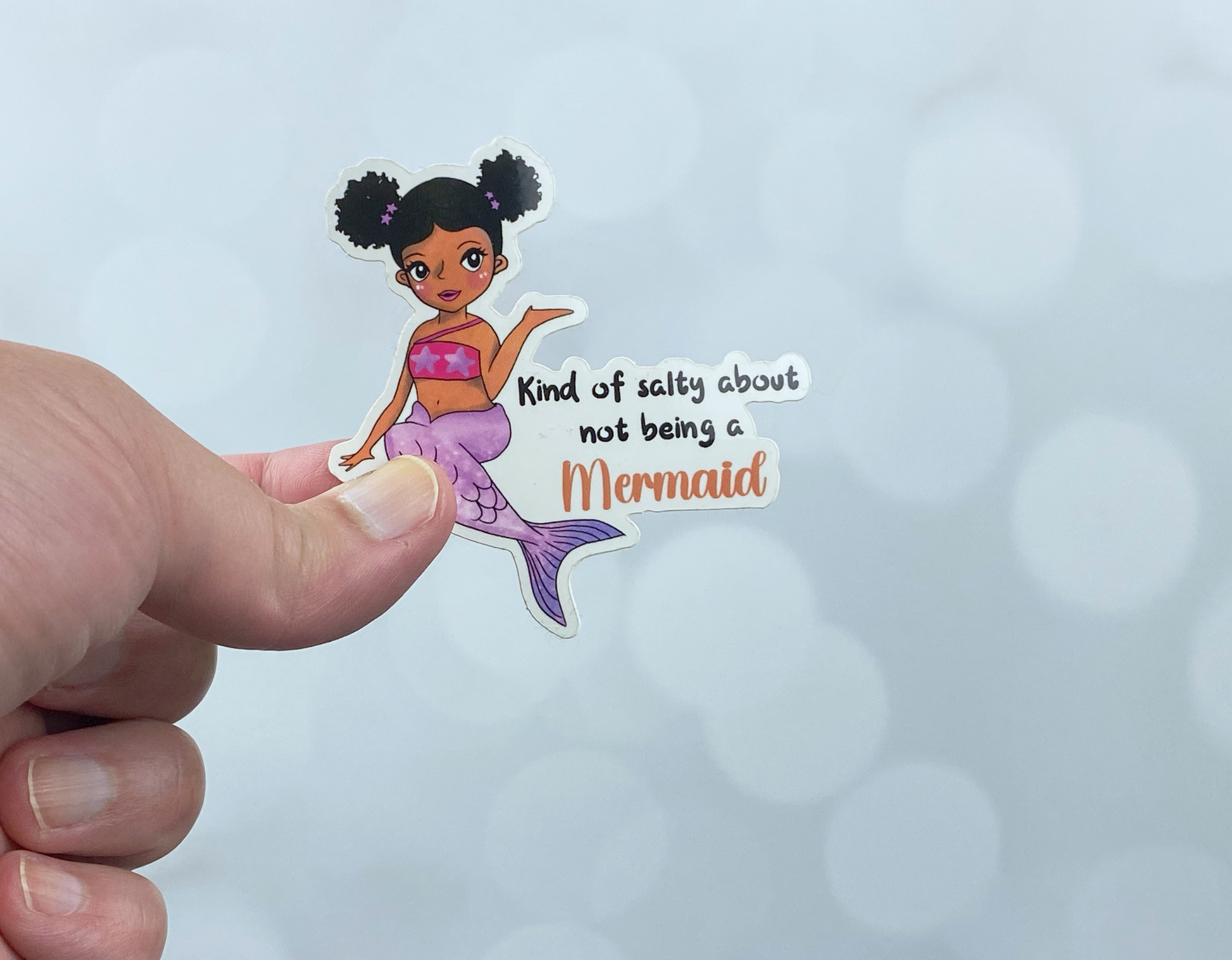 Die-cut vinyl sticker held between fingers, showing a mermaid with black buns and a purple tail, with the text "Kind of salty about not being a mermaid," on a light bokeh background