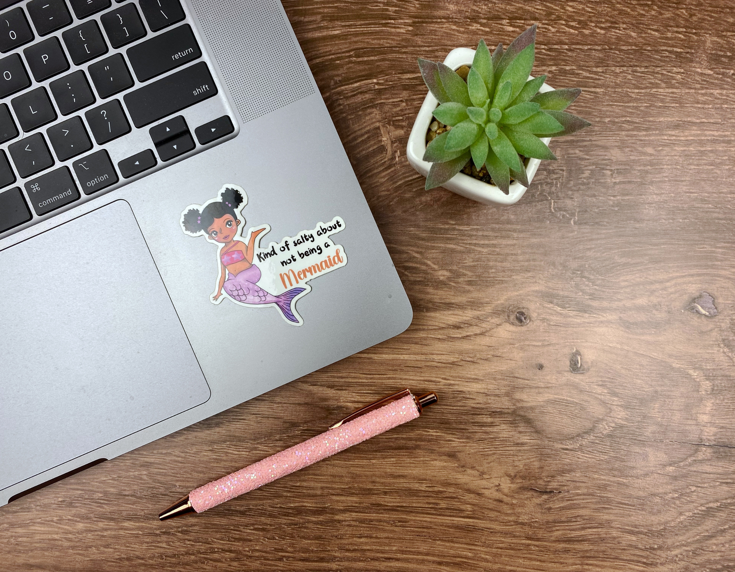 Sassy mermaid sticker applied to the corner of a silver laptop trackpad, next to a succulent and a glitter pen on a wood desk