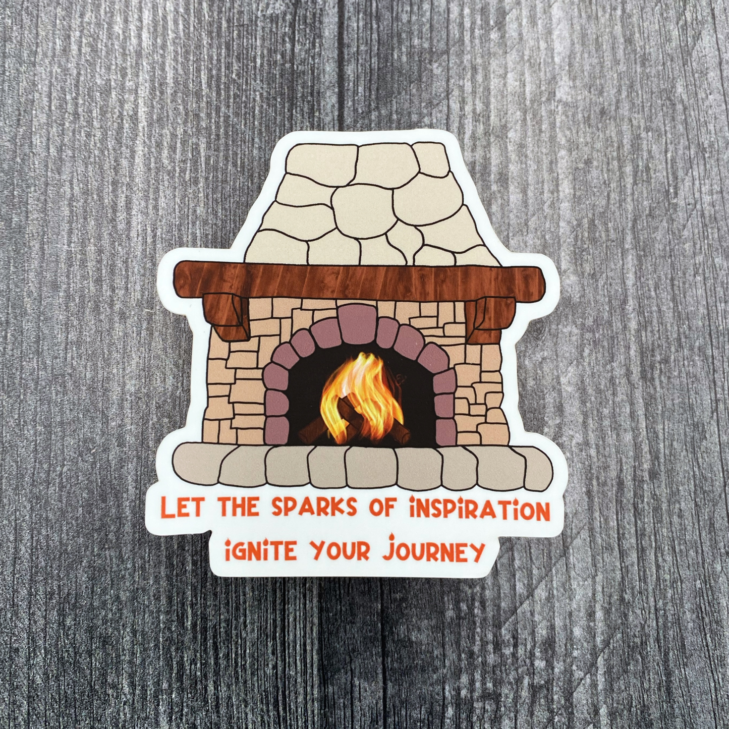 Die-cut vinyl sticker showing a crackling fire inside a stone fireplace, with the text "Let the sparks of inspiration ignite your journey," on a gray wood background