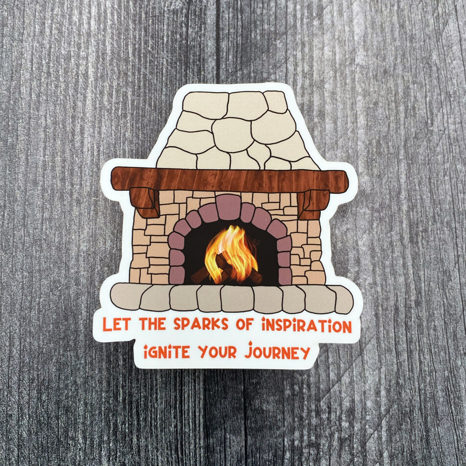 Die-cut vinyl sticker showing a crackling fire inside a stone fireplace, with the text "Let the sparks of inspiration ignite your journey," on a gray wood background
