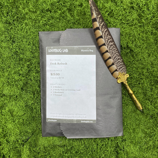 Lootbug Lab Desk Refresh Mystery Bag lying flat in a dark grey kraft paper bag on a vibrant green moss background, with a decorative feather quill pen resting alongside it. A printed label on the bag reads 'Desk Refresh — $23.00' and lists the contents: 2 stickers, 1 sticky note or greeting card, 1 bookmark, and 1 notepad.