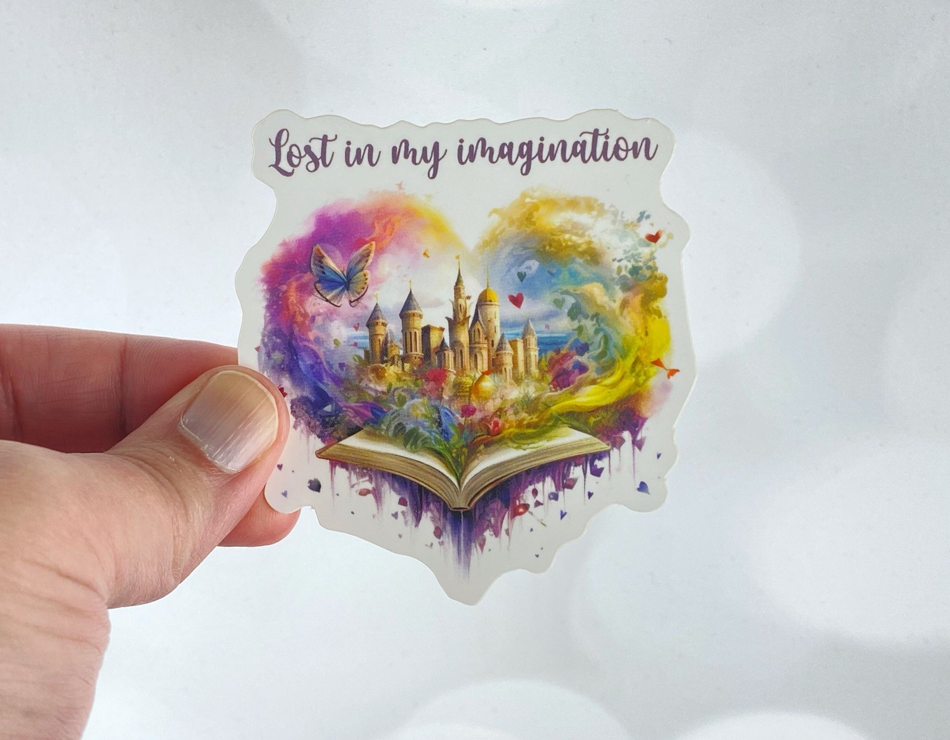 A hand holds a sticker illustration of a fantasy castle blooming from an open book within a heart-shaped watercolor swirl, reading "Lost in my imagination," against a white background.