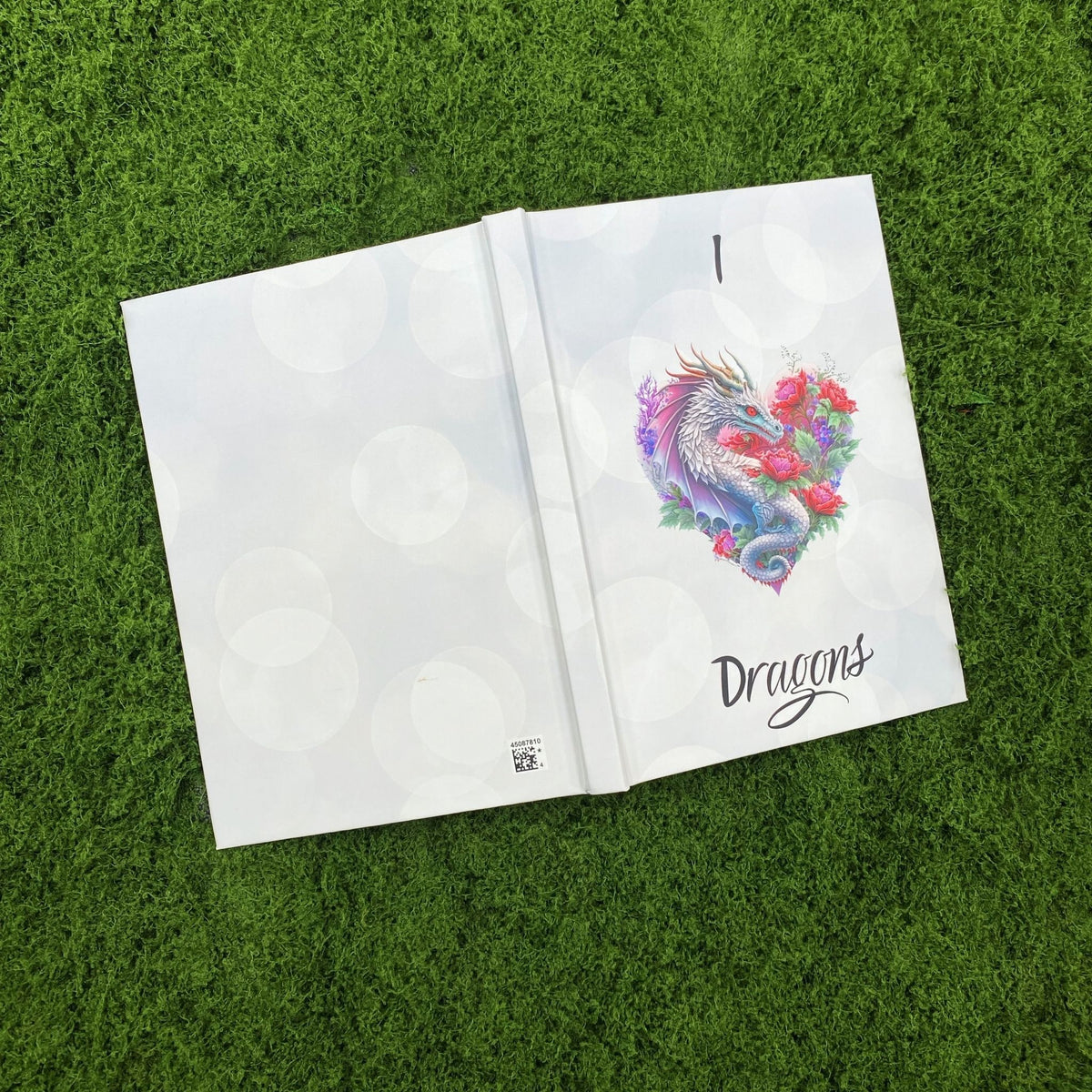 Open white hardcover journal showing the plain bokeh-print back cover and the dragon and rose heart design on the front cover.
