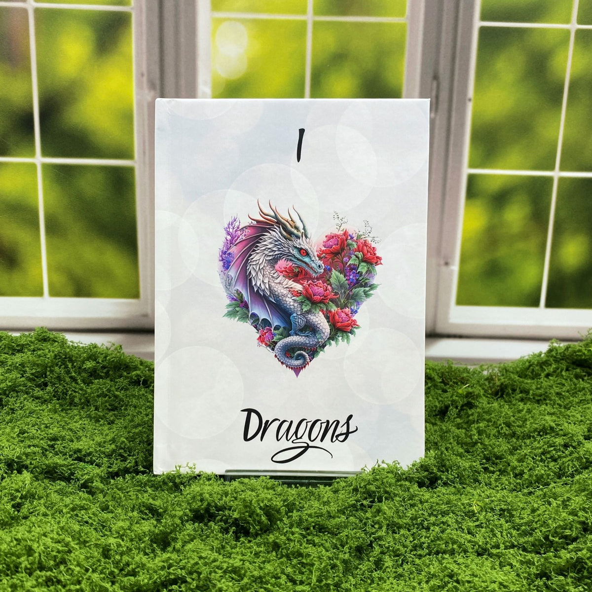 White hardcover journal with a soft bokeh print and a heart-shaped illustration of a dragon wrapped in red roses, reading 'I [dragon heart] Dragons,' displayed on a moss mat with a window backdrop.
