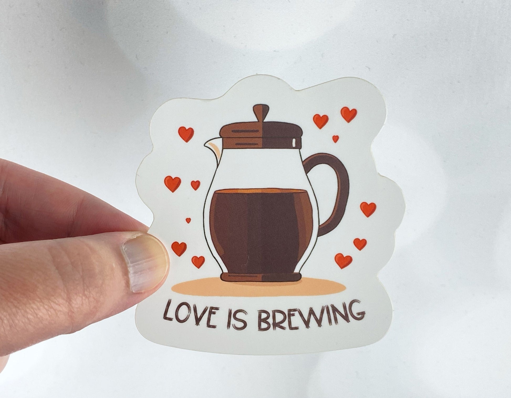 Die-cut vinyl sticker held between fingers, showing a French press full of coffee surrounded by red hearts, above the text "Love is brewing," on a white background
