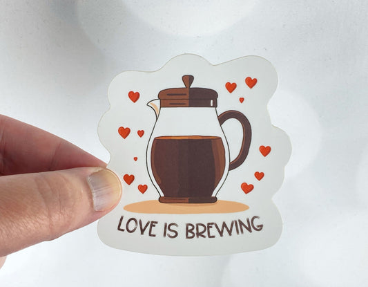 Die-cut vinyl sticker held between fingers, showing a French press full of coffee surrounded by red hearts, above the text "Love is brewing," on a white background