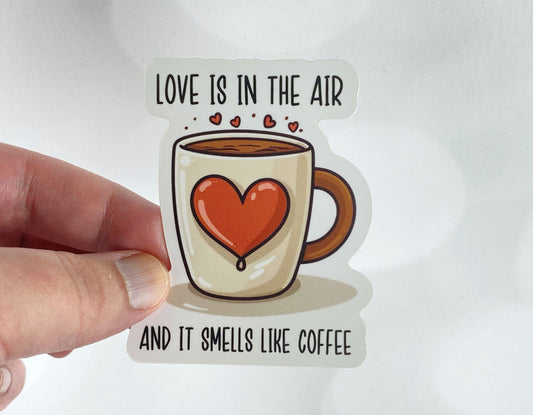 A hand holds a sticker illustration of a coffee mug with a large red heart and steam rising into small hearts, reading "Love is in the air and it smells like coffee," against a white background.