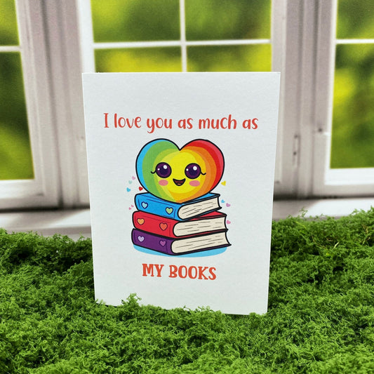 Greeting card that reads "I love you as much as MY BOOKS" above a cartoon rainbow heart character with big eyes sitting on a stack of three books with heart decorations, standing upright on a moss surface in front of a white-framed window.