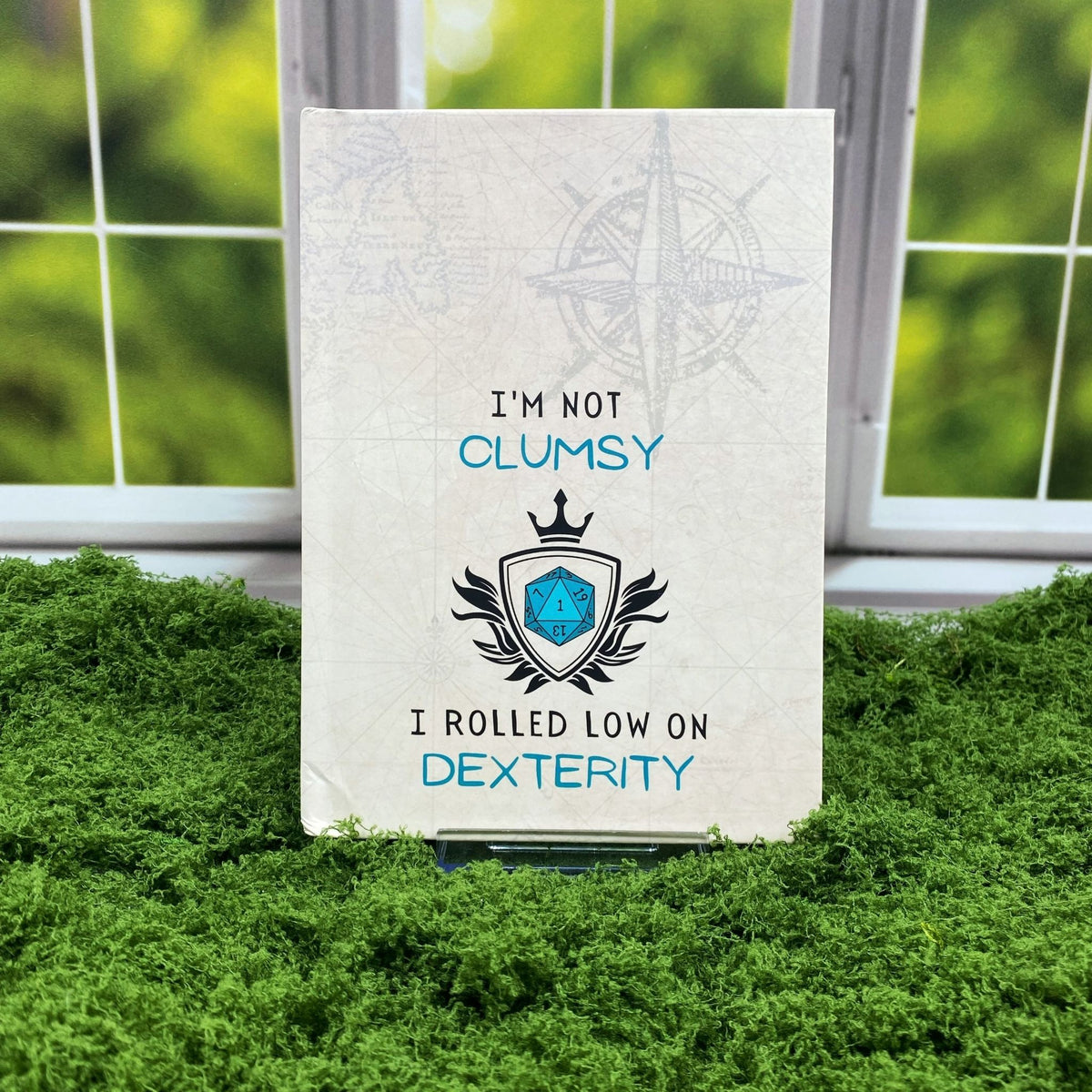 Hardcover journal with a vintage map and compass print, featuring a blue d20 crest and text reading 'I'm not clumsy, I rolled low on dexterity,' displayed on a moss mat with a window backdrop