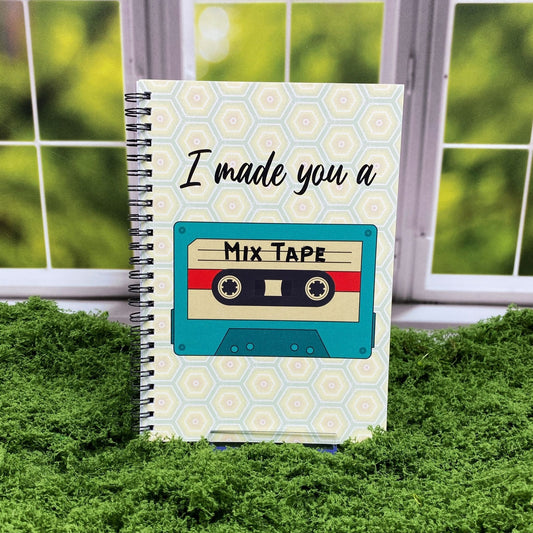 Spiral-bound notebook with a honeycomb pattern cover featuring a teal cassette tape labeled 'Mix Tape' and text reading 'I made you a,' displayed on a moss mat with a window backdrop.