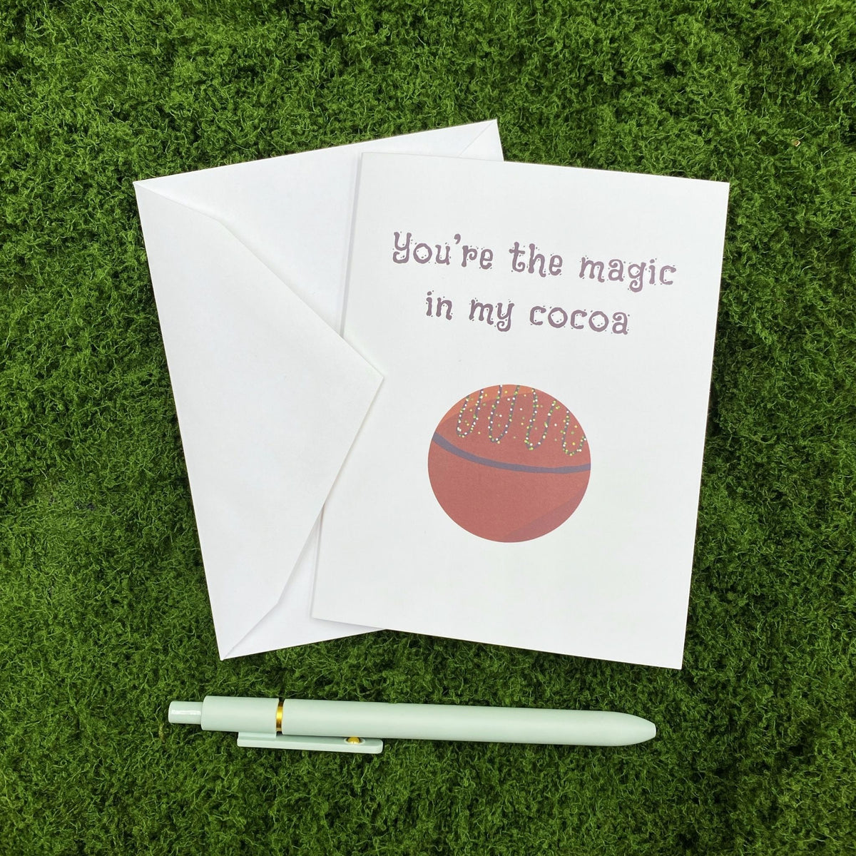 Folded greeting card reading "You're the magic in my cocoa" with a hot cocoa bomb illustration, resting on a white envelope on a mossy surface, with a sage green pen below.