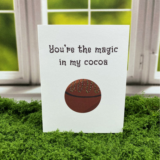 Greeting card that reads "You're the magic in my cocoa" above an illustration of a round hot cocoa bomb drizzled with sprinkles, standing upright on a moss surface in front of a white-framed window.
