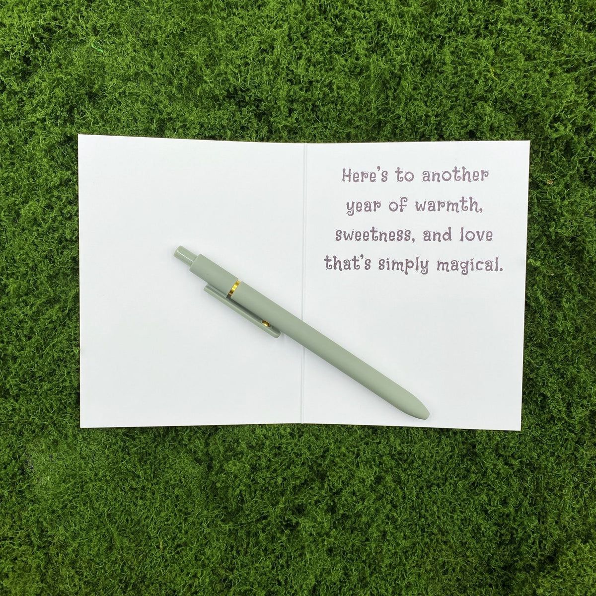 Card opened to show the printed message "Here's to another year of warmth, sweetness, and love that's simply magical" on the right page, with a sage green pen laid across the blank left page on a mossy background.