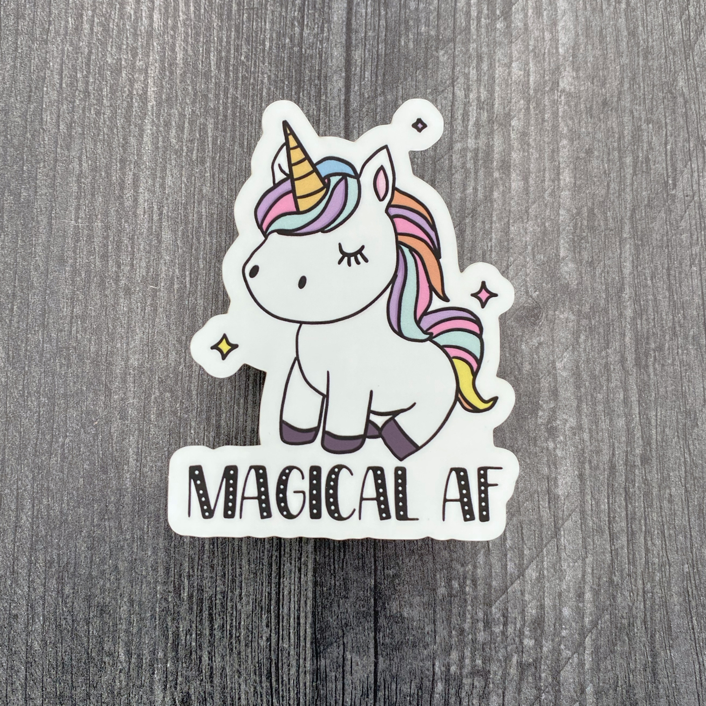 Die-cut vinyl sticker showing a pastel-maned unicorn, with the text "Magical AF," on a gray wood background