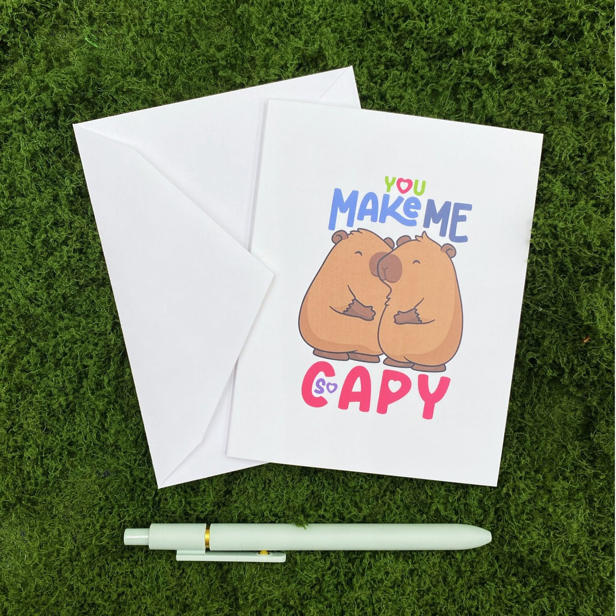 "You Make Me So Capy" greeting card lying on a white envelope on grass, with a mint green pen beside it for scale.