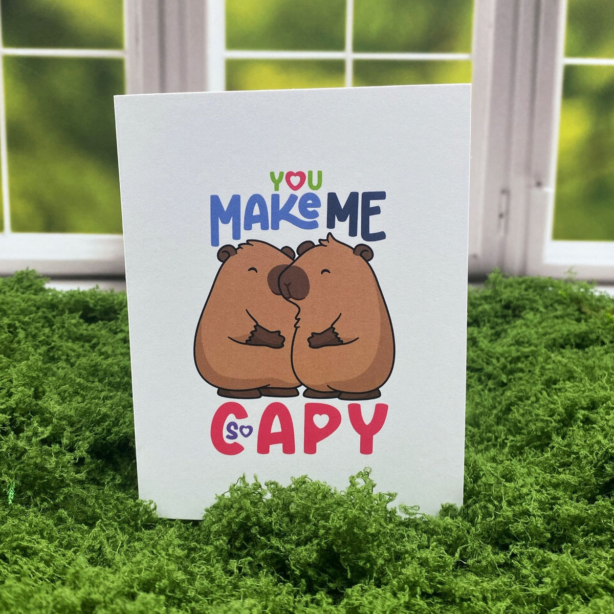 "You Make Me So Capy" greeting card standing upright on a moss mat in front of a window backdrop, featuring two illustrated capybaras nuzzling each other.