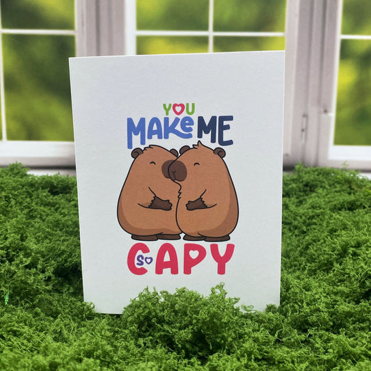 "You Make Me So Capy" greeting card standing upright on a moss mat in front of a window backdrop, featuring two illustrated capybaras nuzzling each other.