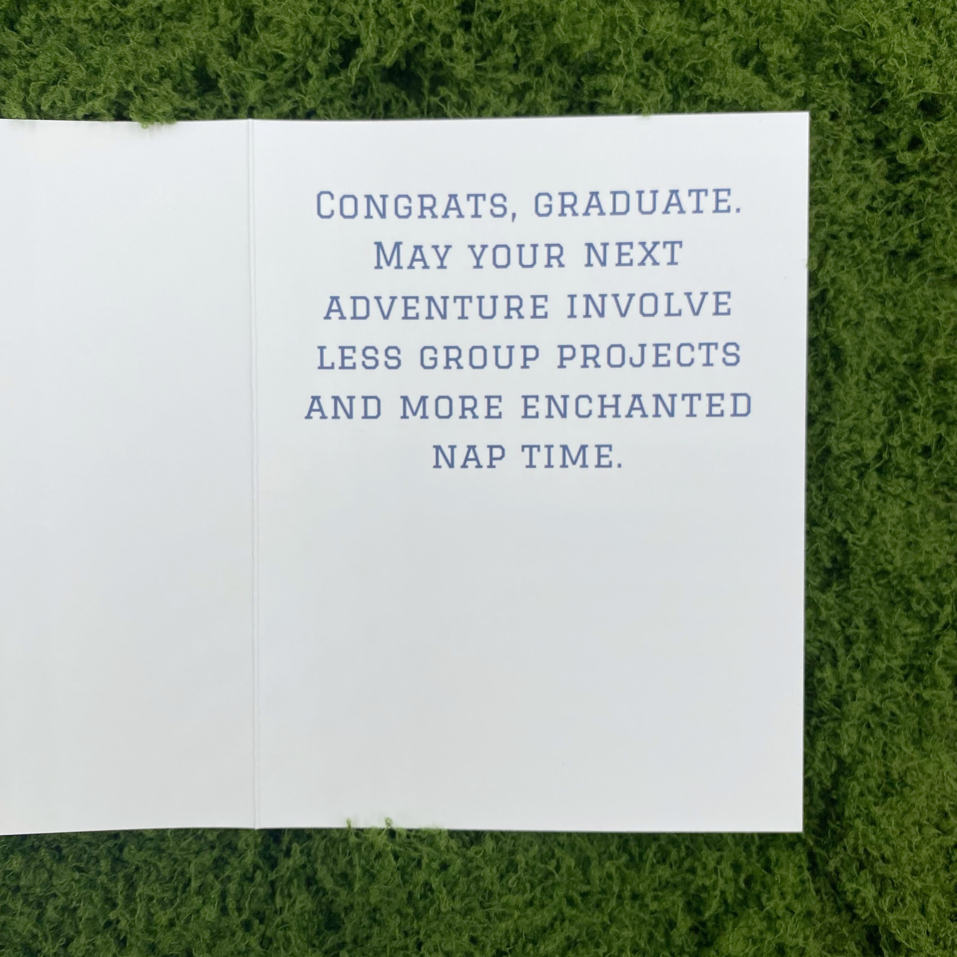 Inside spread of the greeting card showing the printed message "Congrats, graduate. May your next adventure involve less group projects and more enchanted nap time." lying open on artificial grass.