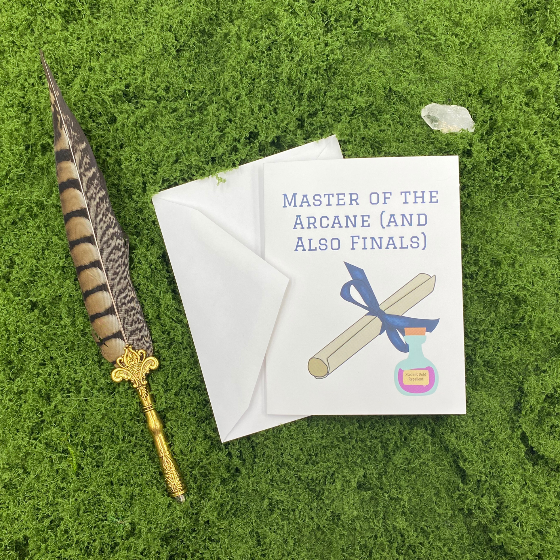 Greeting card that reads "Master of the Arcane (and Also Finals)" with an illustration of a diploma tied in a blue ribbon and a potion bottle labeled "Student Debt Repellent," propped against its envelope on artificial grass with a feather quill pen and a quartz crystal.