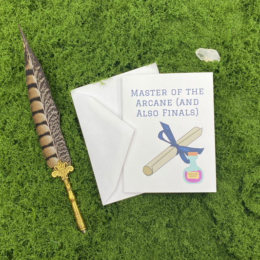 Greeting card that reads "Master of the Arcane (and Also Finals)" with an illustration of a diploma tied in a blue ribbon and a potion bottle labeled "Student Debt Repellent," propped against its envelope on artificial grass with a feather quill pen and a quartz crystal.