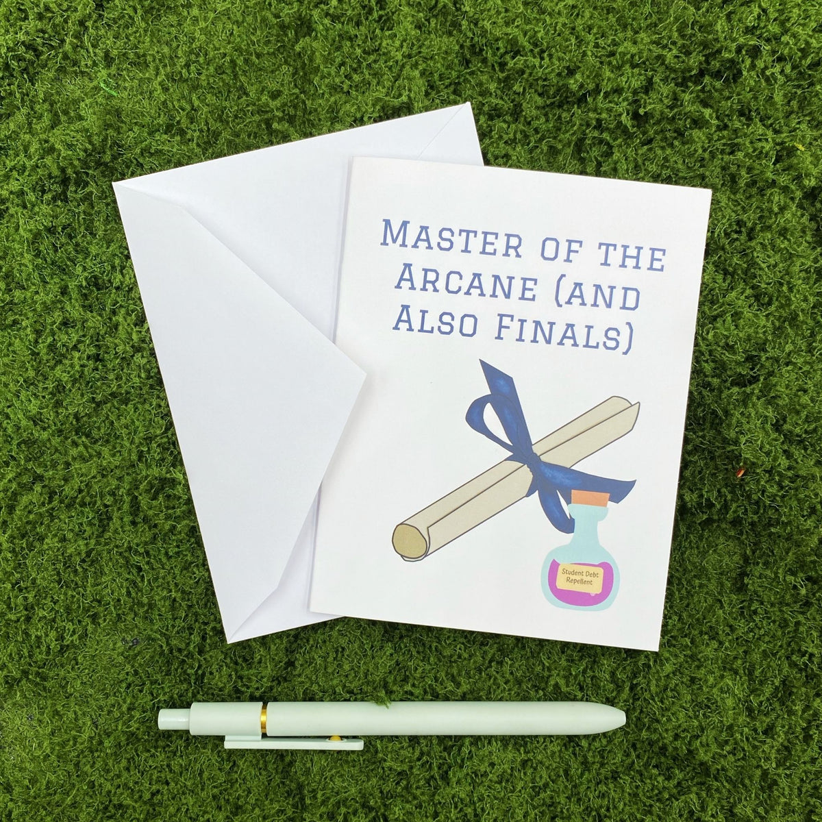 "Master of the Arcane (and Also Finals)" greeting card lying on a white envelope on grass, with a mint green pen beside it for scale.