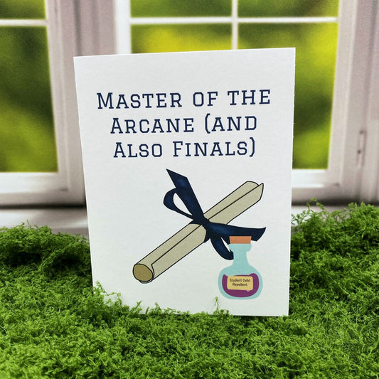 "Master of the Arcane (and Also Finals)" greeting card standing upright on a moss mat in front of a window backdrop, featuring an illustrated diploma tied with a ribbon next to a potion bottle labeled "Student Debt Repellent."
