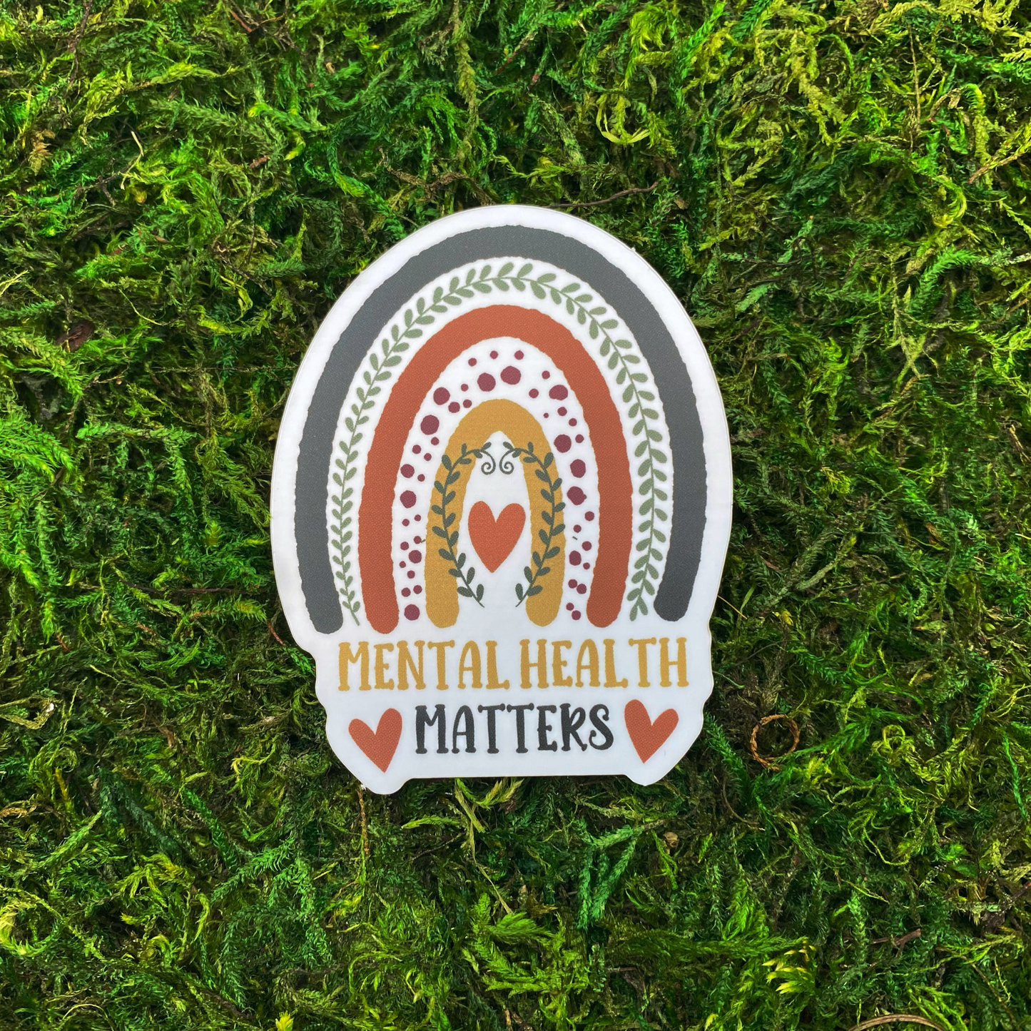 A sticker with an illustration of a boho rainbow decorated with leaf vines and a heart above the text "Mental Health Matters," resting on artificial moss.