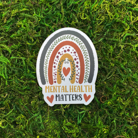A sticker with an illustration of a boho rainbow decorated with leaf vines and a heart above the text "Mental Health Matters," resting on artificial moss.