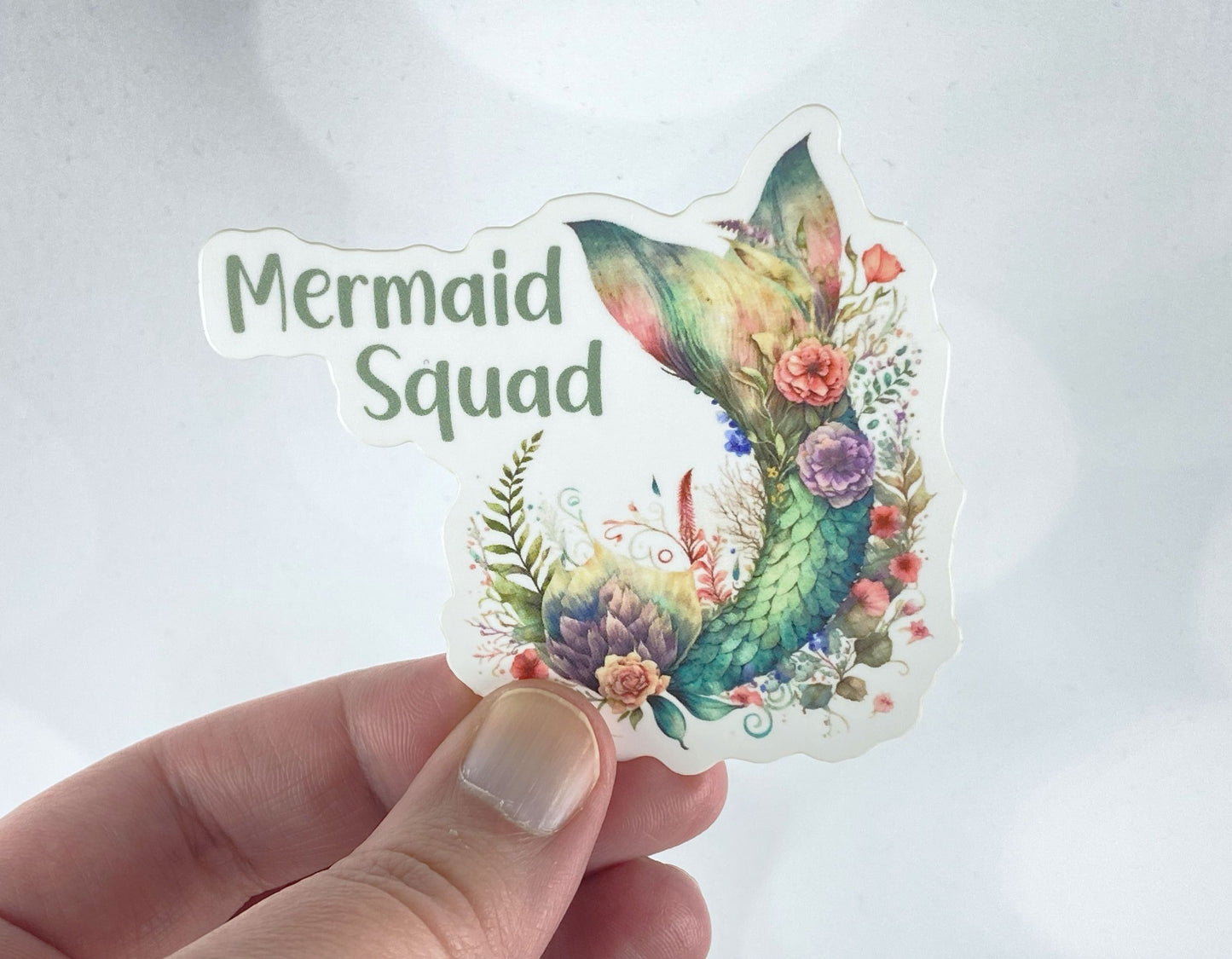 A hand holds a sticker illustration of a jewel-toned mermaid tail surrounded by flowers and greenery, reading "Mermaid Squad," against a white background.